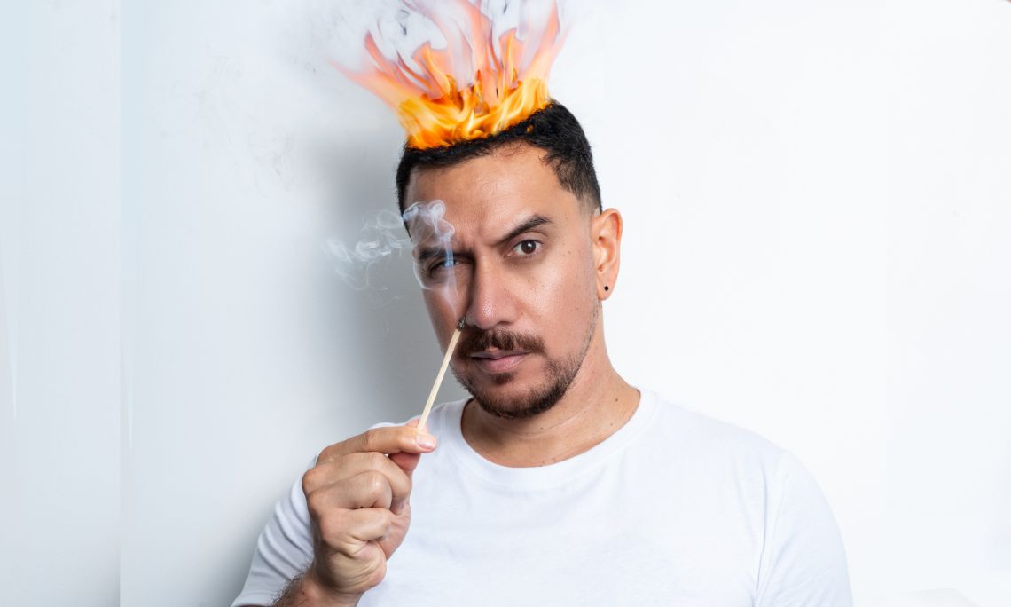 A man looking at camera with eyebrow raised holding a match stick with his hair on fire